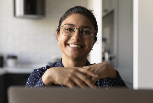 A woman wearing glasses and a navy blue polka dot shirt smiles at the camera while sitting at her desk, hands resting in front of her, with an Infinite RCS platform open on her laptop and a blurred kitchen in the background.