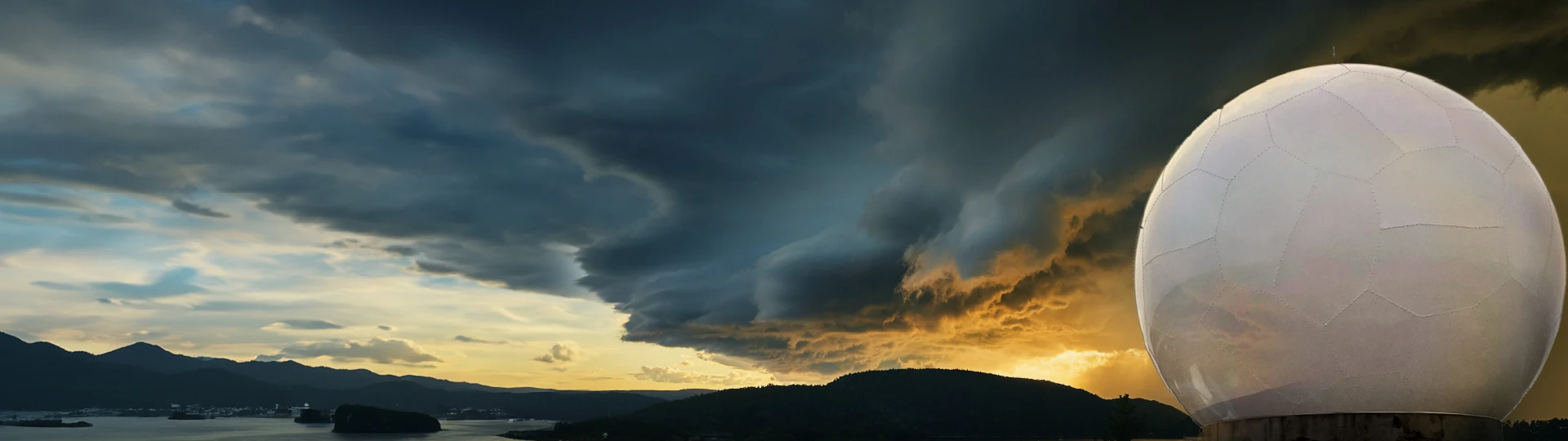 A large white radome stands on a hill at sunset, dramatic clouds above and golden sunlight lighting the landscape and radome below.